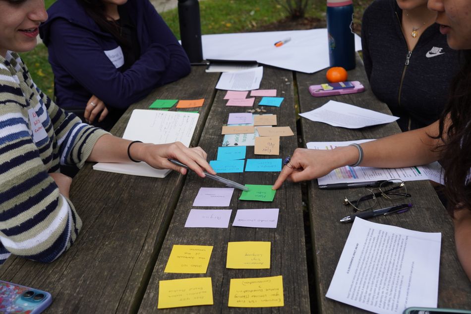 A table with two roys of colorful sticky notes and students pointing at a green sticky note. 