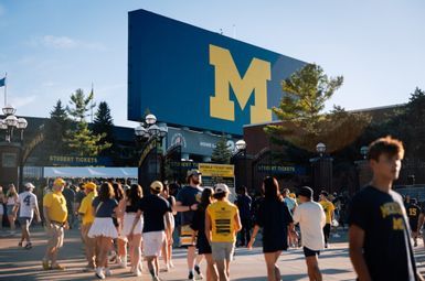 Football fans walk toward the entrance of Michigan Stadium on a clear summer day. A maize block "M" is emblazoned on a blue sign that stands above the crowd.