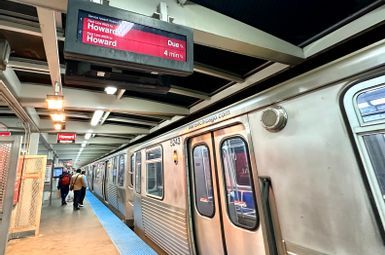 Passengers wait on a platform to board an arriving Chicago Transit Authority light rail train toward Howard Station. An electronic sign shows another is due in 4 minutes.