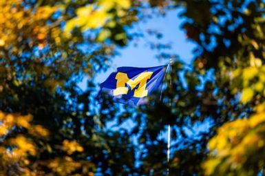 The University of Michigan's maize block M flies on a blue flag, framed by early autumn foliage.