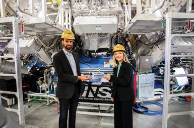 Lawrence Livermore National Laboratory (LLNL) physicists Alex Zylstra and Annie Kritcher stand in the National Ignition Facility (NIF) Target Bay holding a target assembly used in the Aug. 8, 2021 experiment that brought LLNL researchers to the threshold of fusion ignition. Both that experiment and the Dec. 5, 2022 shot that achieved fusion ignition were conducted in the NIF Target Chamber behind them. Photo: Mark Meamber