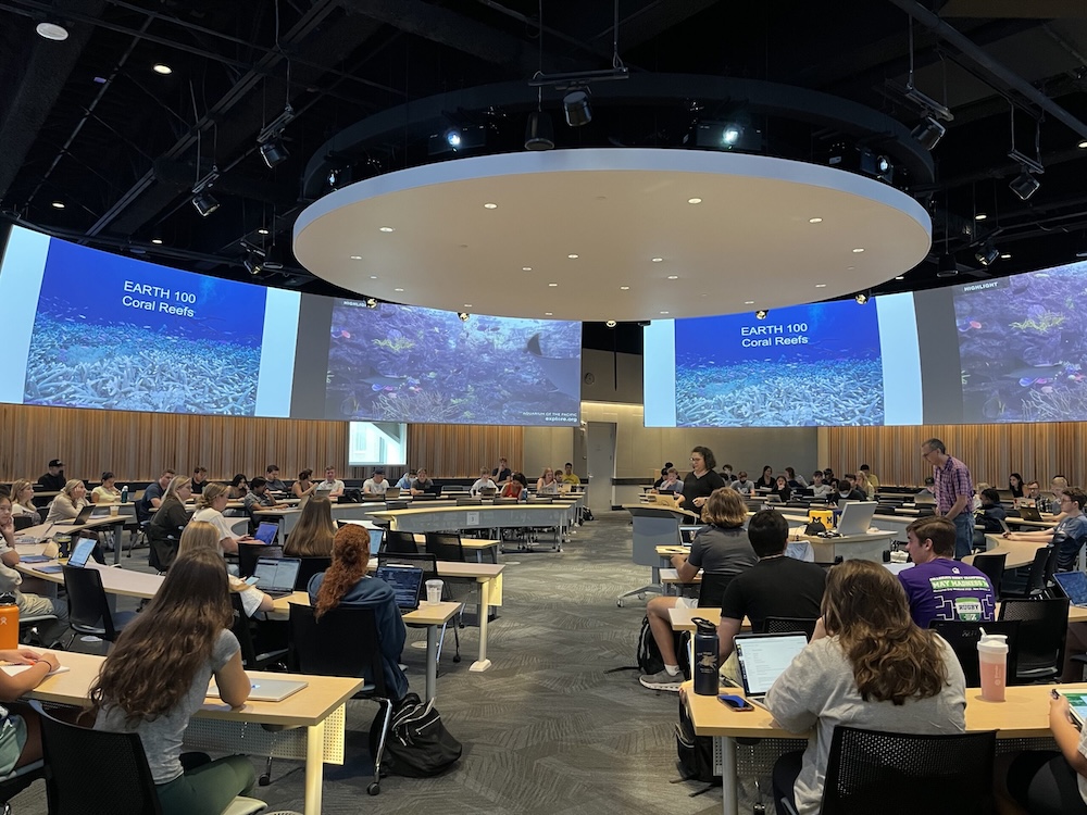 Classroom with students and instructors, large screens displaying "EARTH 100 Coral Reefs."