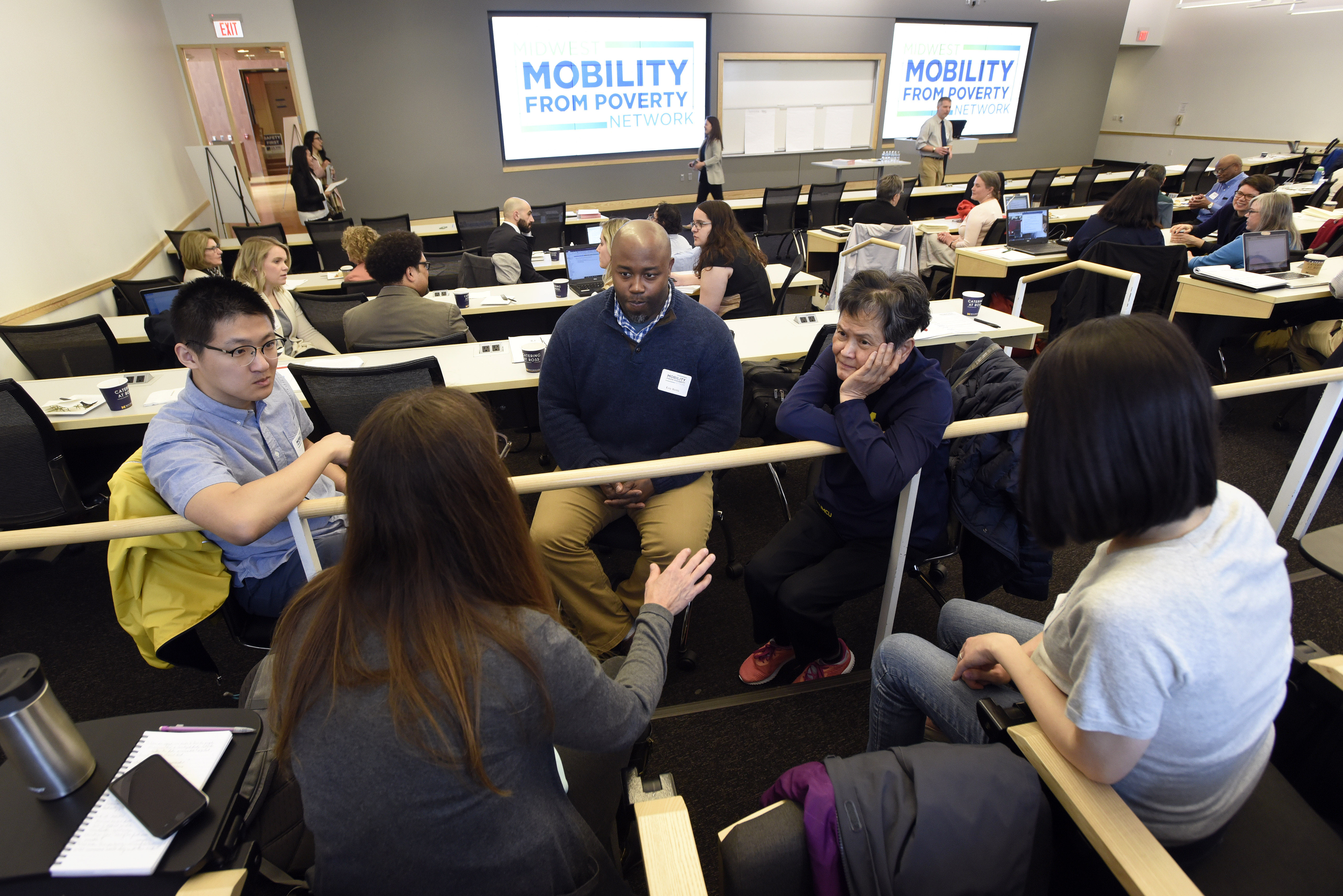 A group of people talking in a meeting room with a slide with the words "Mobility From Poverty Network" projected on screens at the front of the room