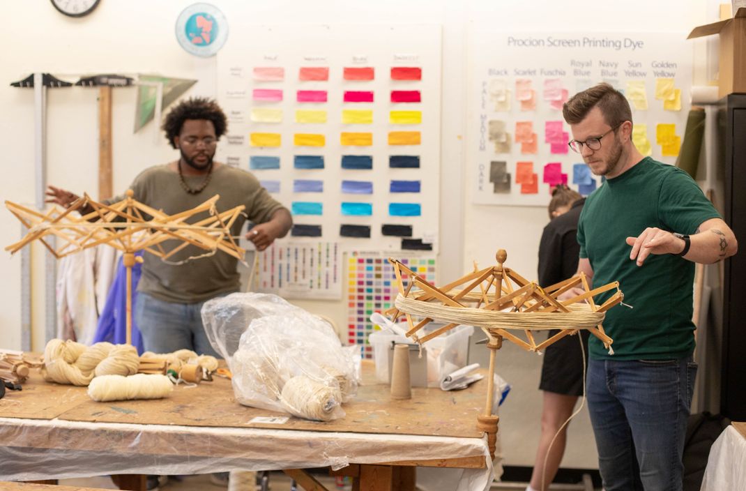 Two people wind yarn onto wooden swifts in a craft room with color swatches on the wall.